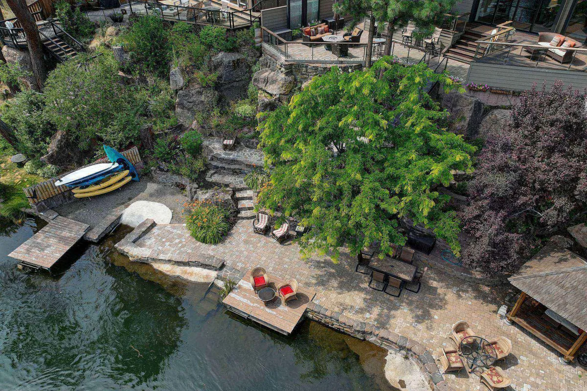 A waterfront seating area beneath a large tree, with kayaks and rock formations nearby, offers a cozy, adventurous vibe. The patio stones and lush greenery set the stage for outdoor dining or peaceful mornings by the water.