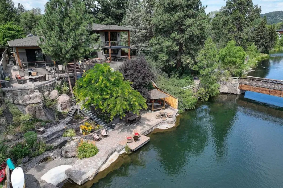 The image showcases a stunning riverfront scene with a spacious deck overlooking the tranquil Deschutes River, surrounded by lush trees. The pathways invite guests down to the water’s edge, where sunset hues reflect off the river, and the outdoor space is perfect for relaxing, enjoying morning coffee, or gathering for an evening bonfire.