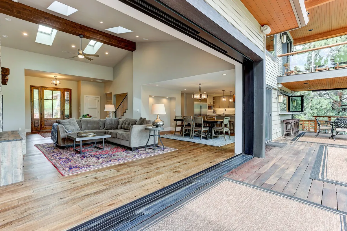 An open-concept interior seamlessly blending living, dining, and kitchen spaces, with a comfortable sofa, a welcoming dining table, a functional kitchen area, and sliding doors leading to an outdoor deck. The room is bathed in natural light, showcasing the effortless flow between spaces and easy access to scenic views.
