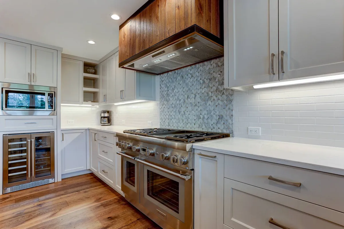 A stylish kitchenette with a stainless steel range, wooden range hood, geometric backsplash, and a wine cooler, designed for both casual snacks and wine tastings.