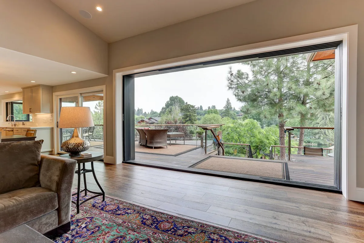 A tranquil interior scene with large sliding glass doors opening to an outdoor deck, a comfortable sofa, an elegant lamp, and a colorful area rug. The view hints at a seamless transition between indoor leisure and outdoor relaxation.