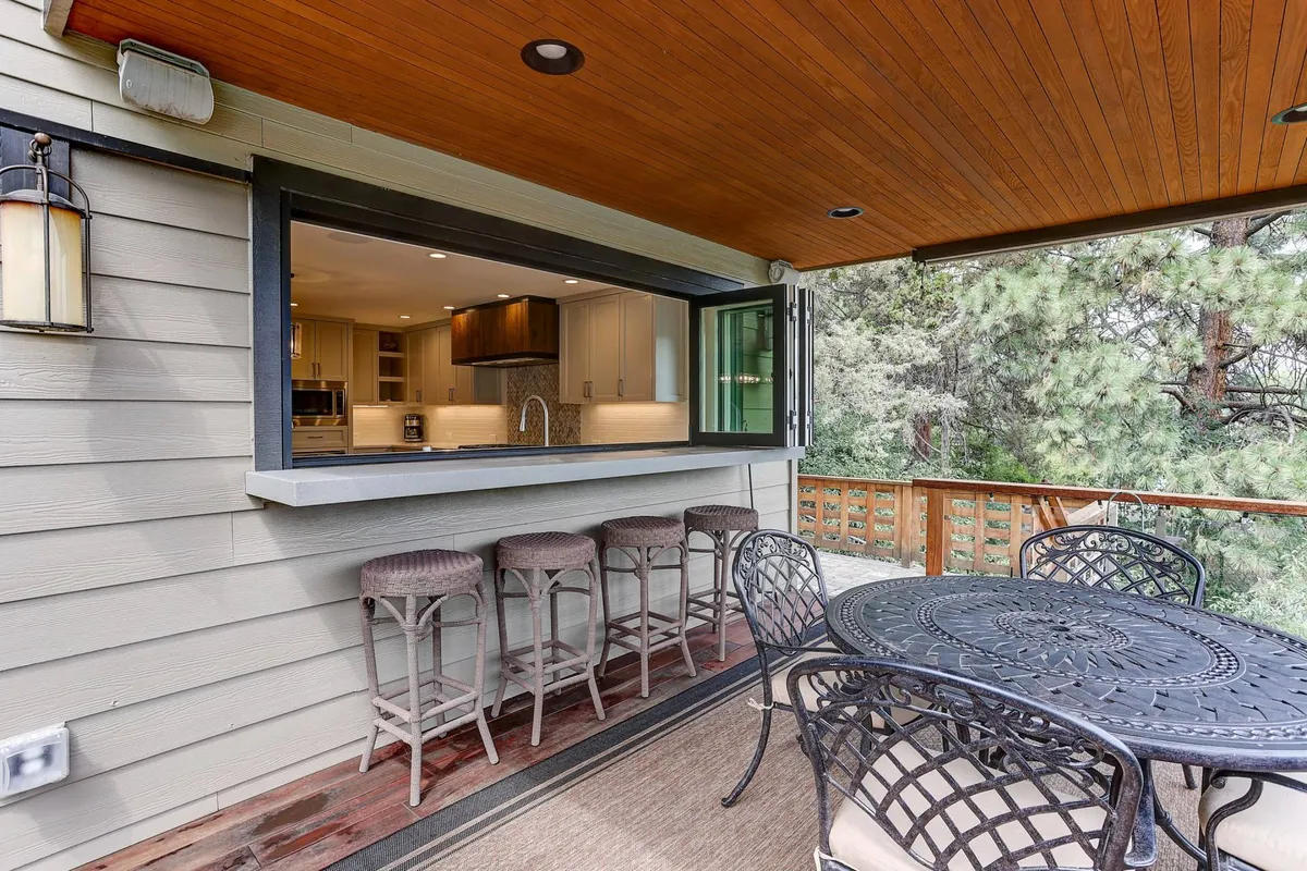 An exterior view featuring an open window connecting the kitchen to an elegantly designed outdoor dining space with stylish bar stools and a large table. The scene suggests seamless indoor-outdoor living, ideal for alfresco meals and social gatherings.