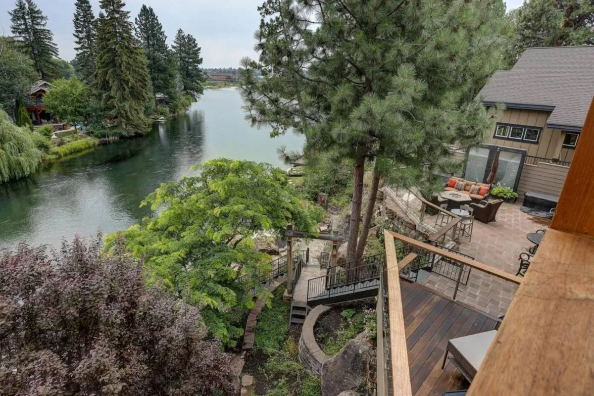 This scenic view captures the tranquil riverfront, an outdoor seating area on a deck, and the lush landscape surrounding the estate. The image highlights the proximity to the river and the peaceful ambiance that guests can enjoy while relaxing, entertaining, or watching children play nearby.