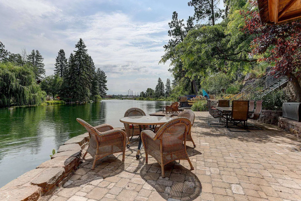 This image features a tranquil lakeside patio with wicker chairs, a stone patio, a table, and surrounding trees. The setting is serene, inviting guests to relax by the water and enjoy the natural beauty, perfect for morning coffee or sunset views.