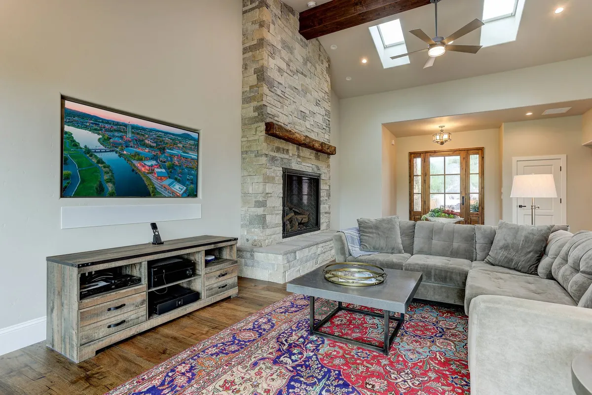 A spacious living room featuring a striking stone fireplace, a large sectional sofa arranged for group gatherings, a central coffee table, and a TV positioned to enjoy both entertainment and the landscape beyond. The room is filled with natural light streaming through expansive windows, while a plush rug and tasteful decor create an inviting atmosphere.