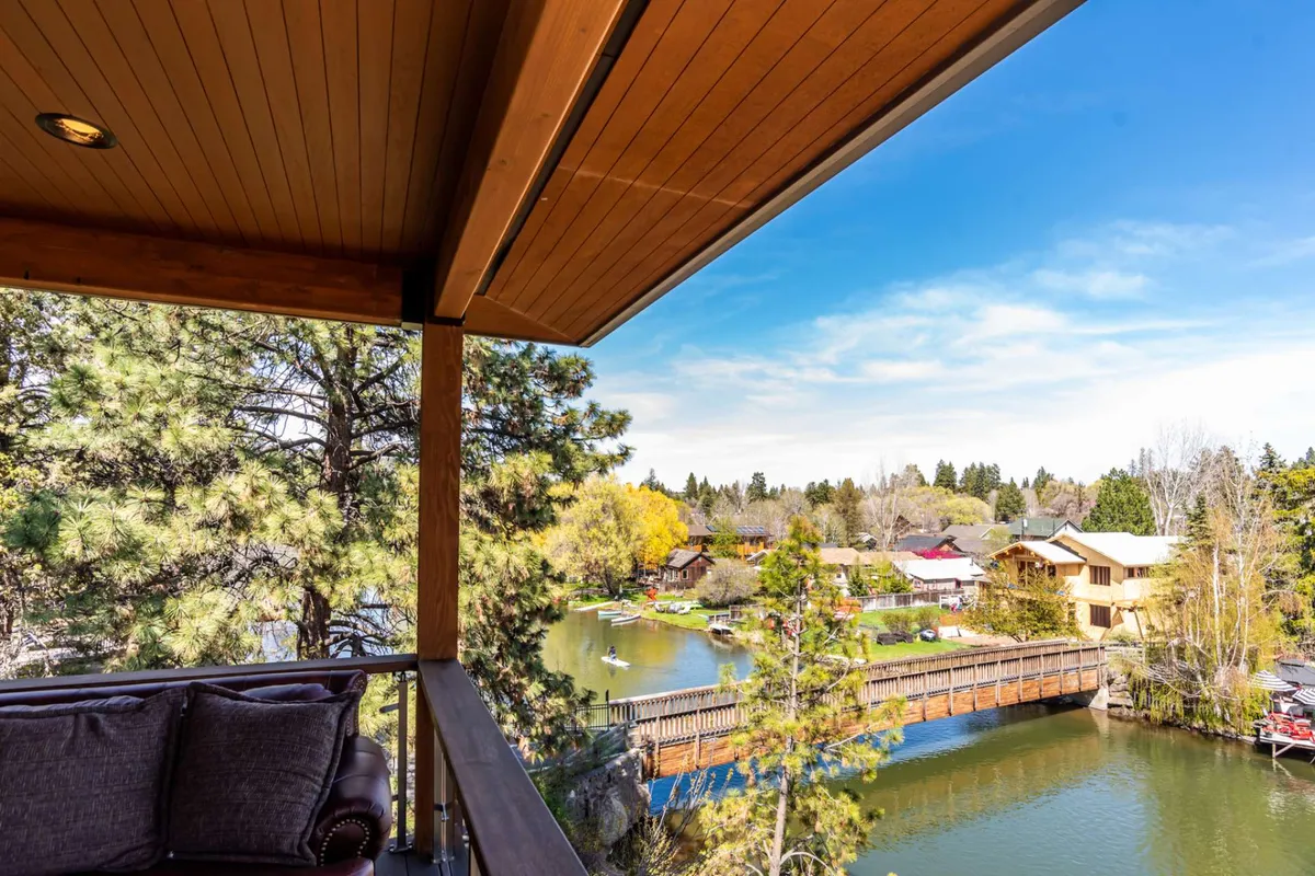 This image features a balcony with comfortable seating overlooking a gentle river, surrounded by leafy trees and a picturesque bridge. In the distance, colorful houses and vibrant community spaces are visible, suggesting proximity to lively neighborhoods and local attractions. The scene embodies tranquility while hinting at the activity and vibrancy nearby.