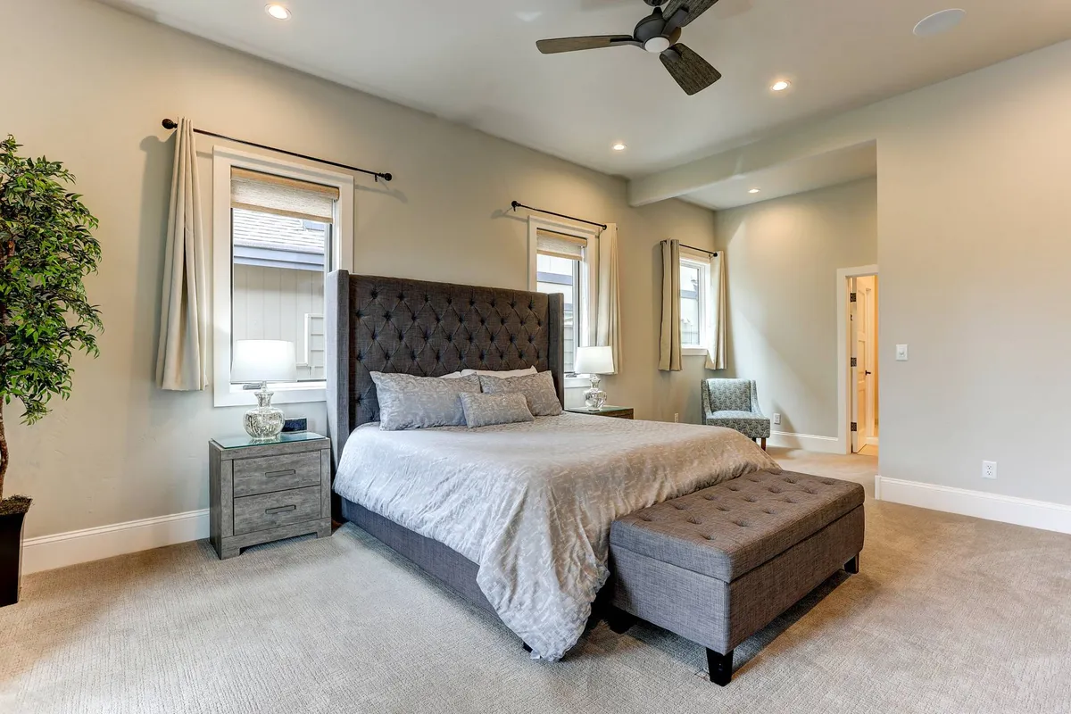 A peaceful upstairs bedroom furnished with a comfortable bed, nightstand, armchair, and decorative plant, illuminated by gentle morning sunlight streaming through windows. The simple, modern style offers a quiet, restful atmosphere.