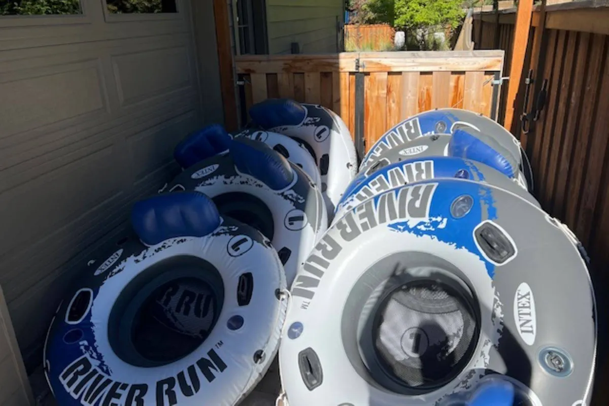 This lively image shows brightly colored inflatable river tubes ready for use, with a wooden fence and garage entrance in the background. The scene evokes the excitement and fun of setting out for a day of river floating with family or friends.