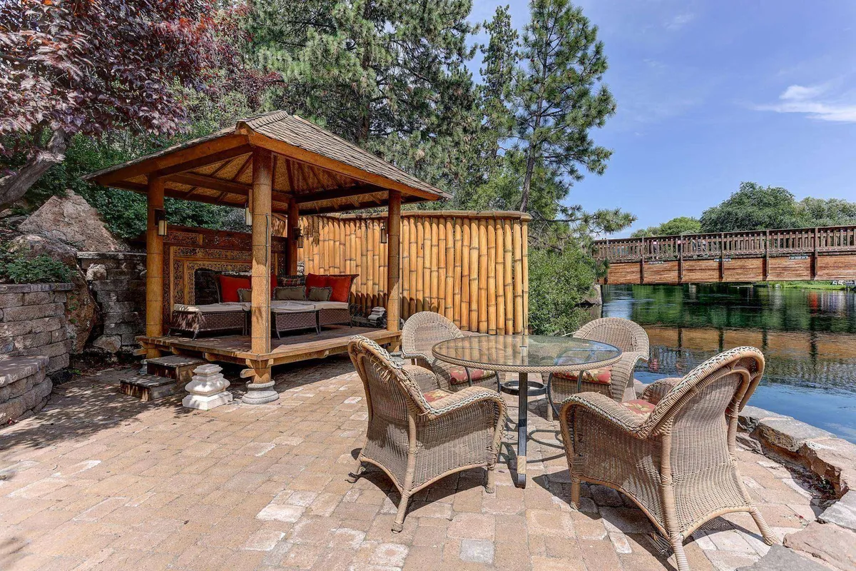 A bamboo gazebo (pagoda) sits beside reflective water, with plush seating and a view of a quaint bridge. The lush landscaping and tranquil setting create a perfect spot for meditation, quiet conversation, or enjoying the sunrise or sunset.