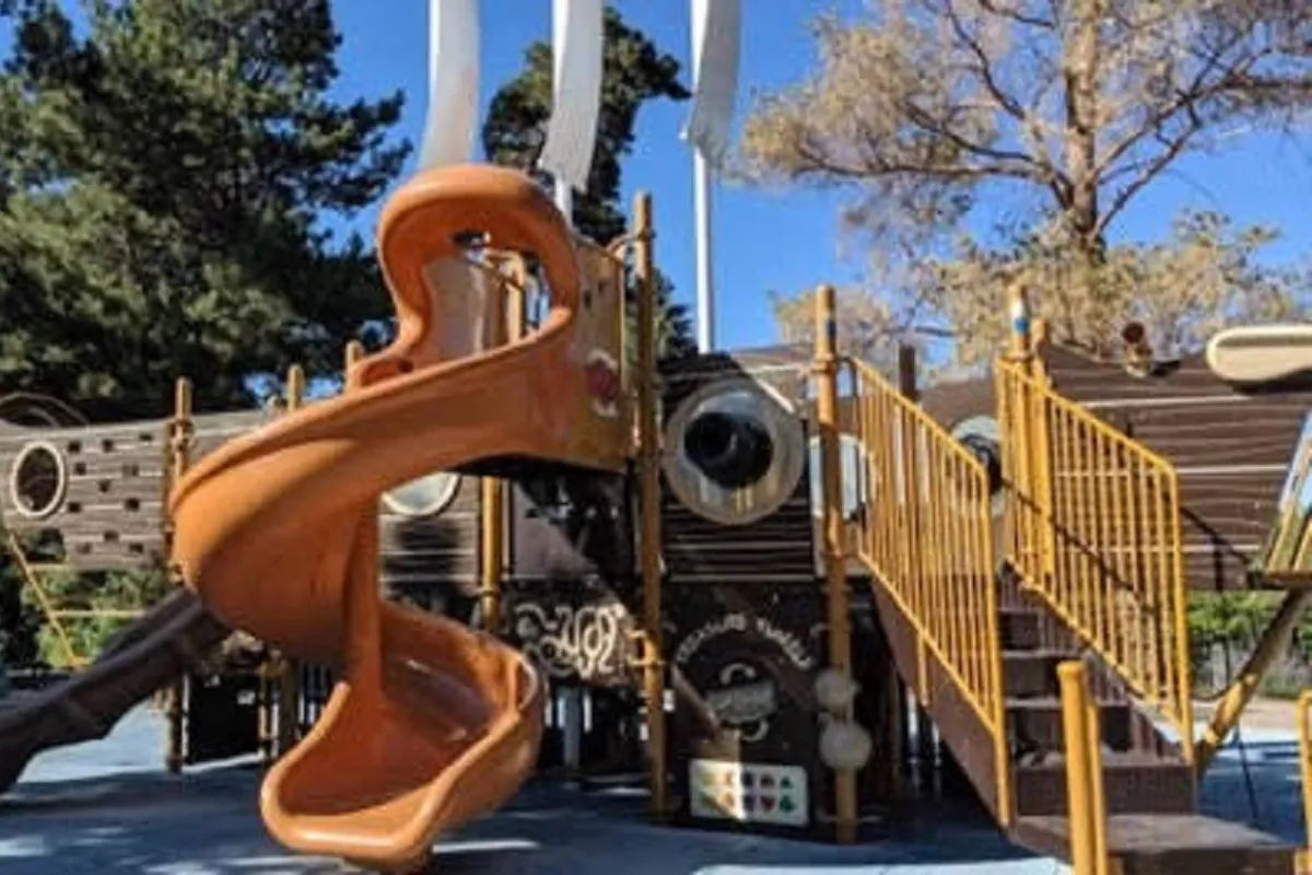 A joyful, nautical-themed playground with an orange slide, climbing structures, and children’s play equipment, all surrounded by nature. The setting is lively and inviting, perfectly suited for family fun and child-friendly adventures.