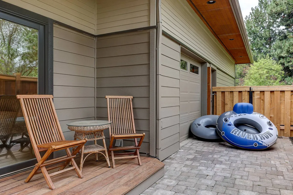 This exterior image features wooden chairs and a small table set on a sunny patio, with inflatable tubes visible, all surrounded by greenery. The relaxed setting invites outdoor lounging and socializing, while the tubes hint at water-based adventures on the nearby river.