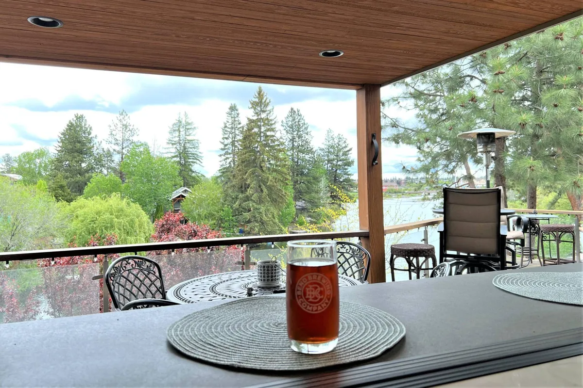 An outdoor seating area with a table and a glass of drink overlooks a lake framed by trees. The mood is peaceful and inviting, suggesting gatherings with friends or quiet moments spent observing nature and enjoying the views.