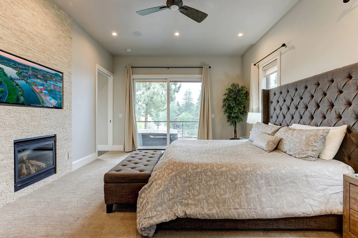 A spacious bedroom filled with natural light, featuring a large bed with a tufted headboard, a fireplace, wall-mounted TV, and floor-to-ceiling windows looking out to lush greenery. Stylish modern décor and an indoor plant add a calming, luxurious ambiance.