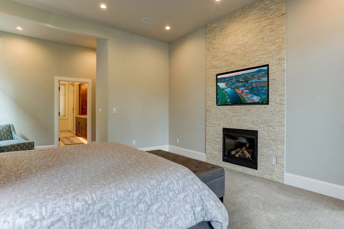 A serene bedroom with a large bed, fireplace, and a wall-mounted TV, all highlighted by soft, natural light. The setting exudes comfort and calm, inviting restful sleep and relaxation.